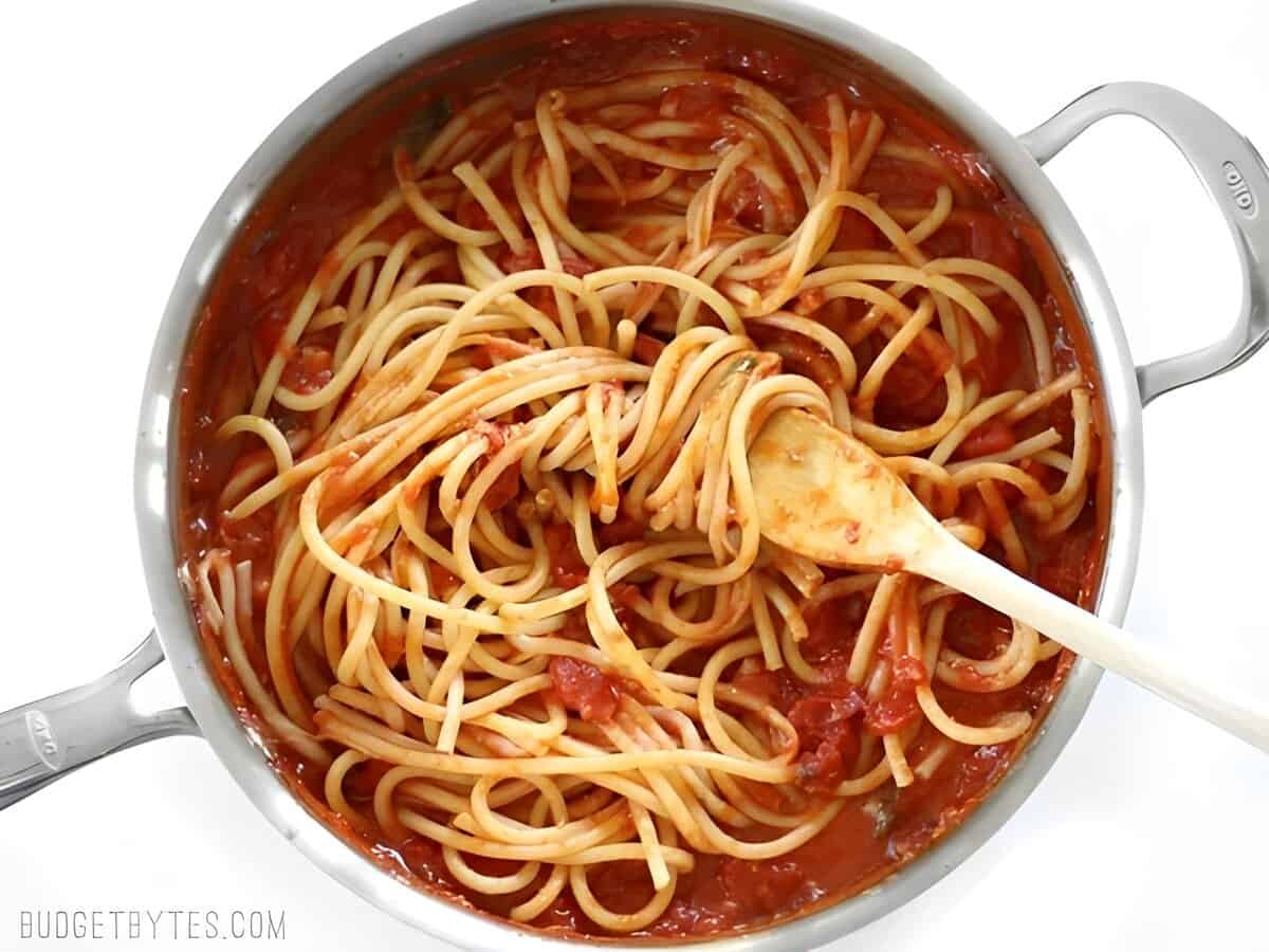 Spaghetti added to homemade tomato sauce in a skillet with a wooden spatula.