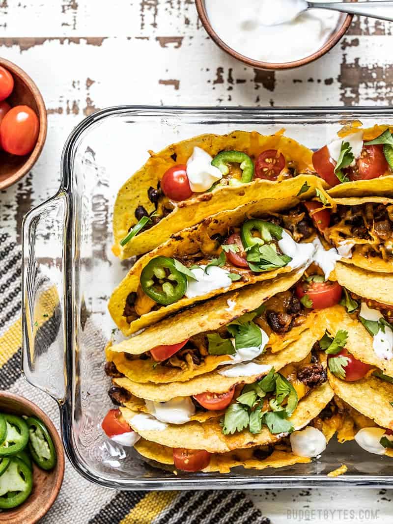 Baked Beef and Black Bean Tacos are a fast and easy way to take Taco Tuesday to the next level and is the perfect fast and easy weeknight meal. Budgetbytes.com A glass casserole dish filled with baked beef and black bean tacos, with bowls of toppings on the sides