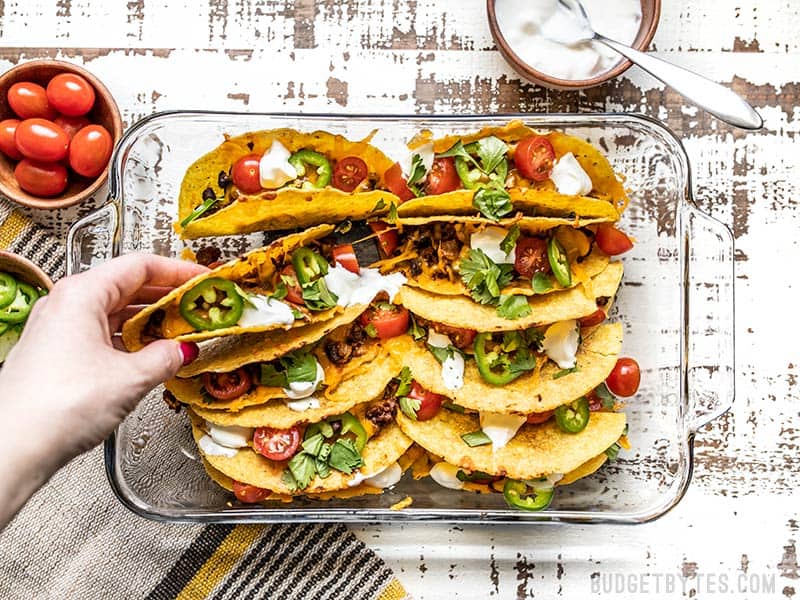 Baked Beef and Black Bean Tacos are a fast and easy way to take Taco Tuesday to the next level and is the perfect fast and easy weeknight meal. Budgetbytes.com A hand picking up one of the baked beef and black bean tacos out of the casserole dish