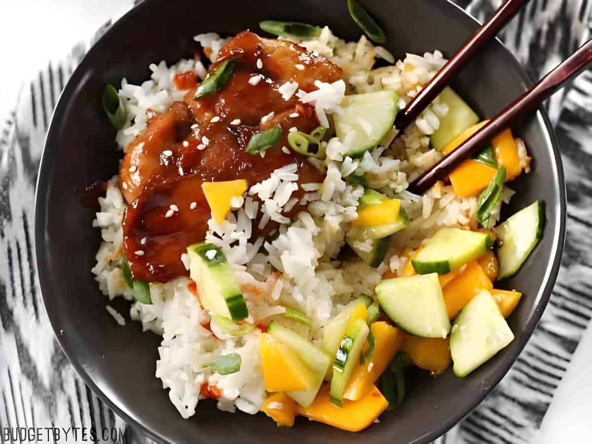Soy ginger chicken in bowl with white rice, mango, and cucumber slices.
