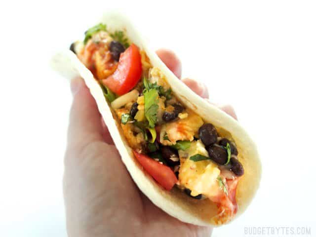 Ultimate Southwest Scrambled Eggs make a fast and filling dinner or brunch, and are a great way to use up leftover ingredients in the kitchen. BudgetBytes.com A hand holding a taco filled with Ultimate Southwest Scrambled Eggs