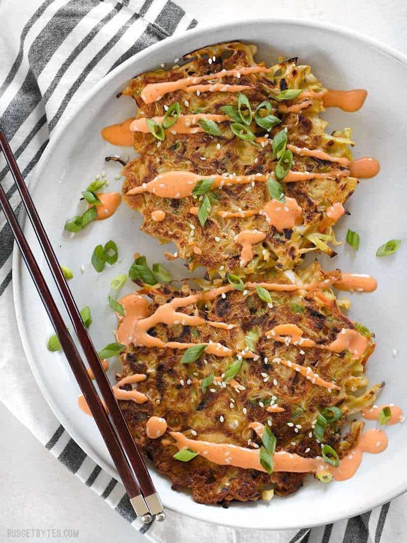 Savory Cabbage Pancakes are a fun and filling way to use up pantry leftovers. Fill them and top them with whatever your heart desires! BudgetBytes.com Two Savory Cabbage Pancakes on a plate drizzled with sriracha Mayo, green onion, and sesame seeds.