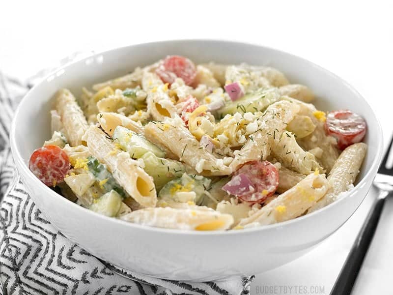 Creamy Lemon Dill Greek Pasta Salad is packed with bold flavors and fresh vegetables, making it a delicious light lunch or cold summer meal. BudgetBytes.com Side view of Creamy Lemon Dill Greek Pasta Salad in a bowl on a striped napkin with a black fork on the side