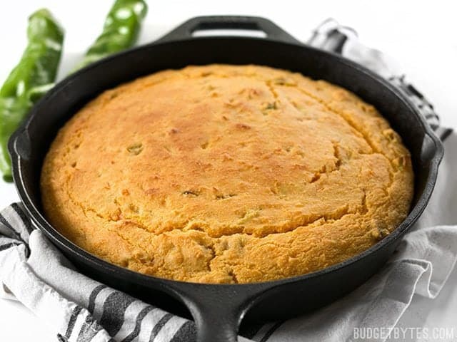 Side view of Hatch Chile Sweet Potato Cornbread in cast iron skillet sitting on napkin with two chilis in the background