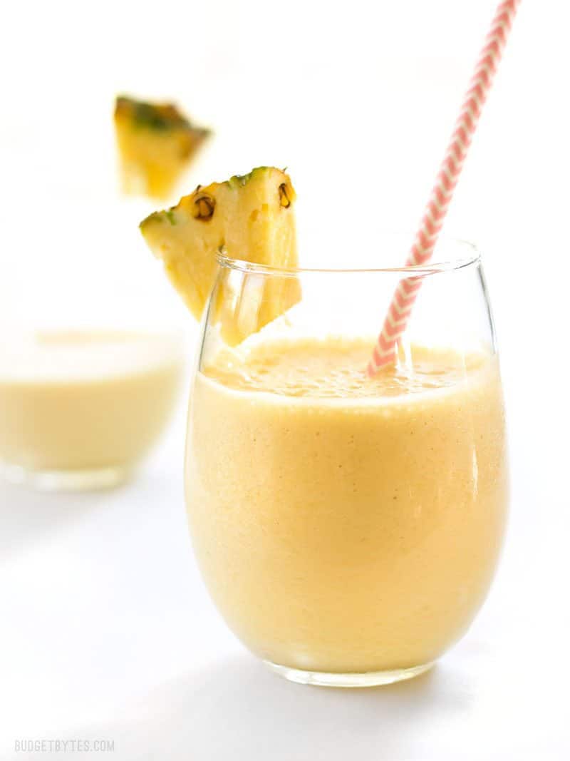 When you need a tropical escape this homemade Pineapple Orange Julius is the perfect sweet and creamy frozen drink to take you away. BudgetBytes.com Two glasses of homemade Pineapple Orange Julius with pineapple slices on the rims and pink paper straws