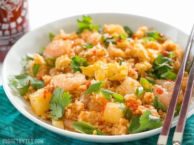 Shrimp Fried Rice with Pineapple and Toasted Coconut is a fast and easy alternative to takeout, perfect for busy weeknights! BudgetBytes.com Front view of a bowl of Shrimp Fried Rice with Pineapple and Toasted Coconut with a bottle of sriracha in the back.