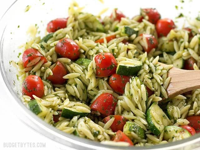 Zucchini and Orzo Salad with Chimichurri is a fresh summer salad that makes the perfect fresh and zesty side dish for summer grilling. BudgetBytes.com Close up side view of the bowl full of Zucchini and Orzo Salad with Chimichurri, with a wooden spoon stuck in the side