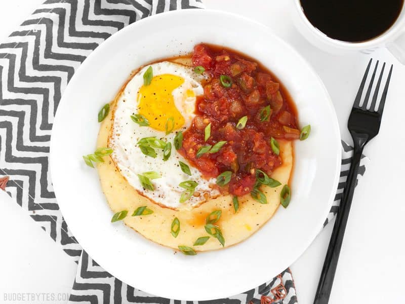 Cheddar Grits Breakfast Bowls are an easy but indulgent breakfast or brunch treat that you can whip up in no time. BudgetBytes.com Overhead shot of a Cheddar Grits Breakfast Bowl on a striped napkin with coffee nearby