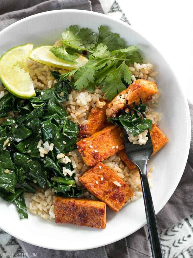 Chili Garlic Tofu Bowls are a flavor filled healthy lunch that you can pre-pack for your week ahead. Easy Vegan Meal Prep. Close up of Chili Garlic Tofu Bowls with a fork piercing a piece of tofu and some kale.