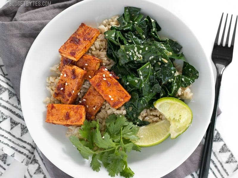 Chili Garlic Tofu Bowls are a flavor filled healthy lunch that you can pre-pack for your week ahead. Easy Vegan Meal Prep. BudgetBytes.com A Chili Garlic Tofu Bowl garnished with lime wedges and cilantro, a fork on the side.