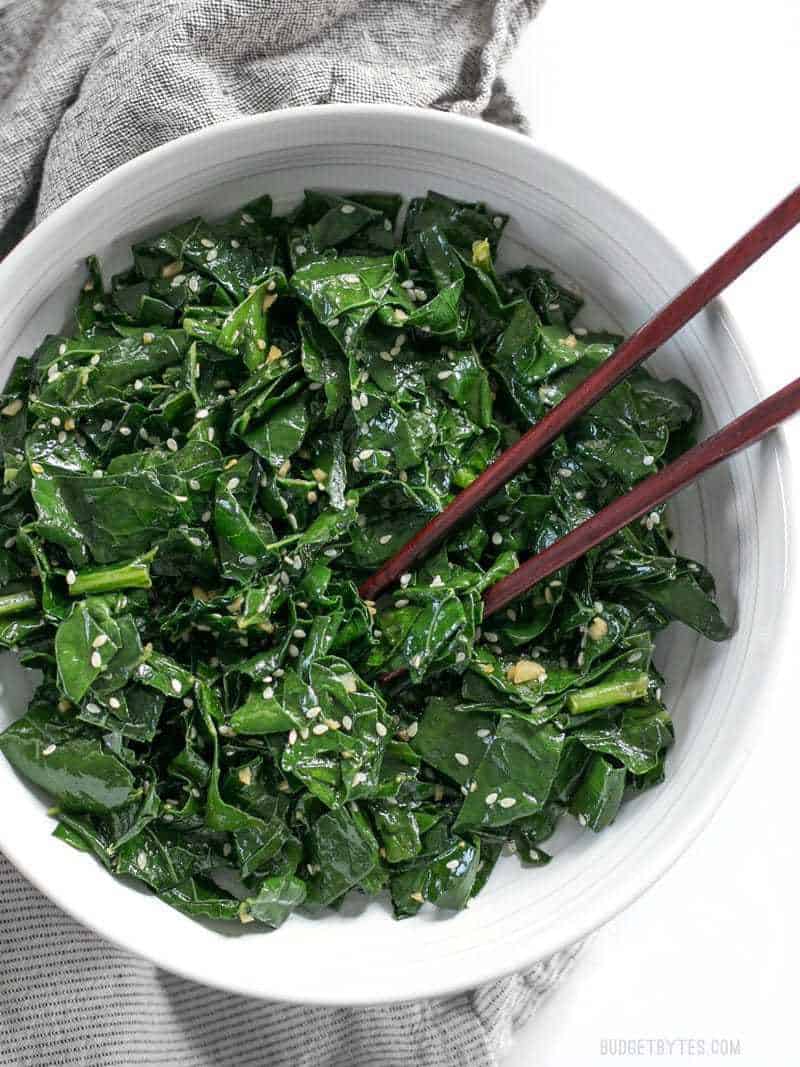 Sesame Kale is a quick, easy, and satisfying side dish that pairs perfectly with any Asian inspired meal. BudgetBytes.com A big bowl of Sesame Kale with wooden chopsticks about to pick up a piece.