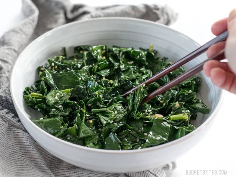 Sesame Kale is a quick, easy, and satisfying side dish that pairs perfectly with any Asian inspired meal. BudgetBytes.com Front view of Sesame Kale being picked up with chopsticks out of the bowl