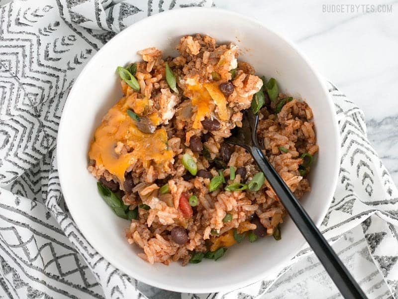 This Beef Burrito Casserole is totally customizable and can include all your favorite burrito or nacho toppings. Leftovers reheat great for lunch! Budgetbytes.com A bowl of Beef Burrito Casserole being eaten with a black fork, on a black and white patterned napkin.