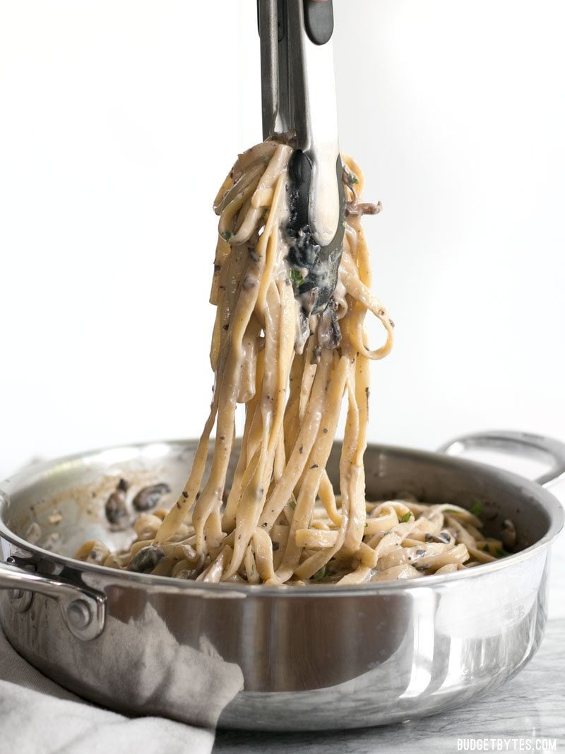 This rich and Creamy Mushroom Herb Pasta is a surprisingly fast and simple to way to have a gourmet meal at home. BudgetBytes.com Tongs lifting a bunch of Creamy Mushroom Herb Pasta out of the skillet, viewed from the side.
