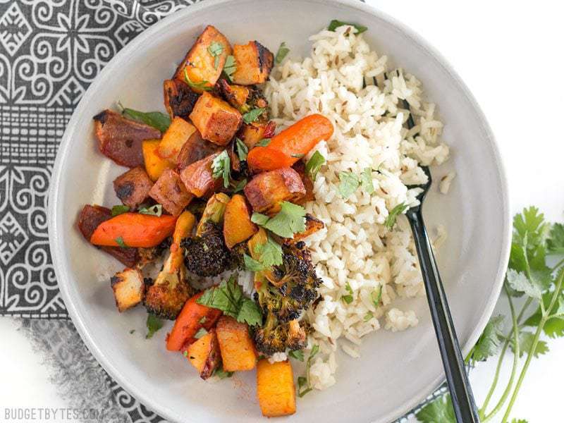 Harissa Roasted Vegetables are a spicy, slightly sweet, and smoky vegetable medley that makes a great side for meat or fish. BudgetBytes.com Harissa Roasted Vegetables in a bowl with cumin rice, garnished with cilantro