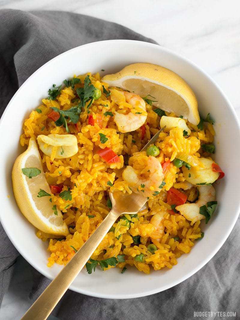 Seafood Rice Skillet is a nod to seafood paella using easy to find ingredients and equipment. Impress your dinner guests with this easy and impressive dish! BudgetBytes.com Close up of a bowl of Seafood Rice Skillet with a gold fork scooping a bite.
