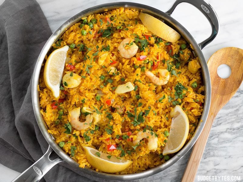 Seafood Rice Skillet is a nod to seafood paella using easy to find ingredients and equipment. Impress your dinner guests with this easy and impressive dish! BudgetBytes.com Overhead view of the Seafood Rice Skillet garnished with lemon wedges and parsley