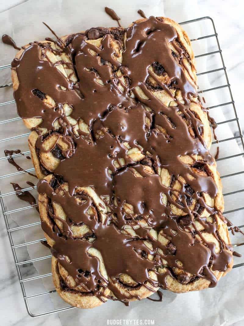 A 5 ingredient dough makes these chocolate cinnamon buns fast, easy, soft, and fluffy. The perfect treat for lazy weekend mornings! BudgetBytes.com 12 chocolate cinnamon buns drizzled with chocolate icing, cooling on a wire cooling rack
