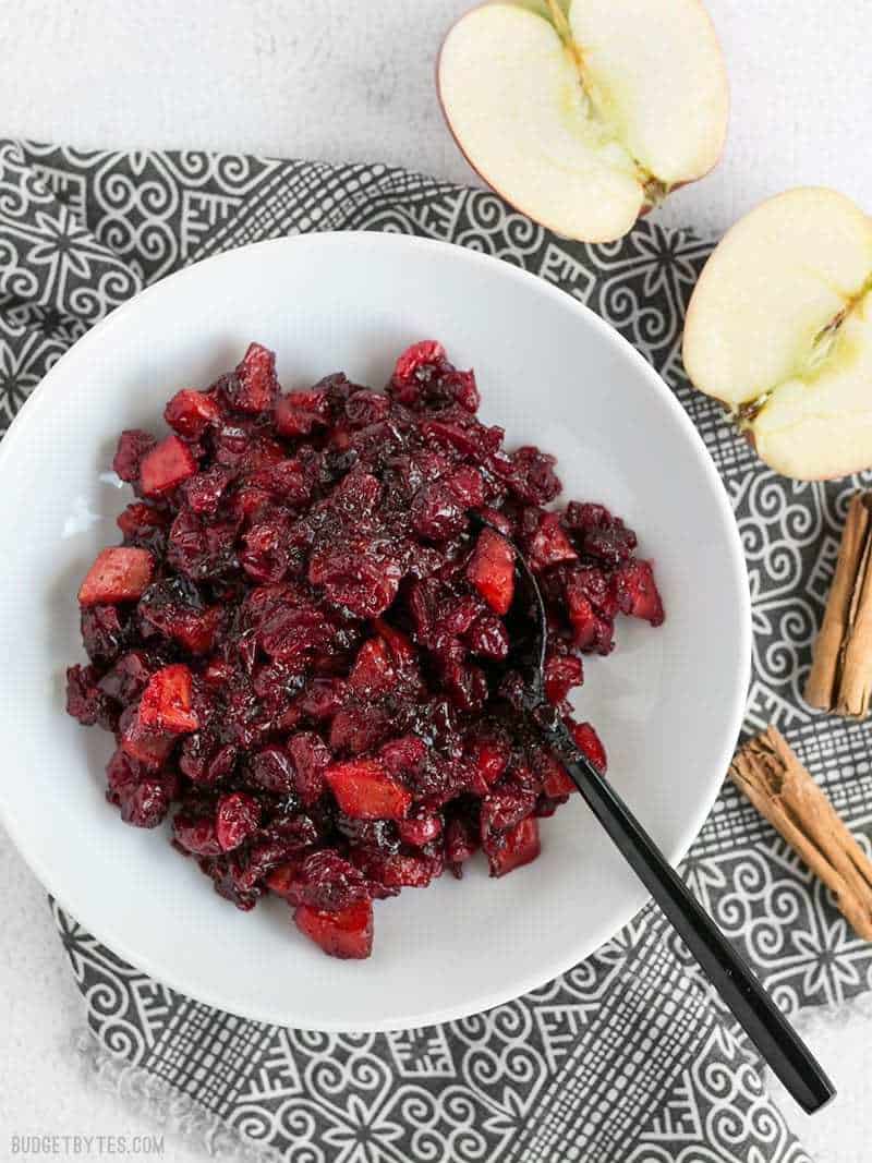 Roasted Apple Cranberry Relish is a thick, jam-like condiment whose uses go far beyond the Thanksgiving table. BudgetBytes.com Roasted Apple Cranberry Relish in a white bowl with a black fork, on a patterned napkin, apples and cinnamon sticks on the side.