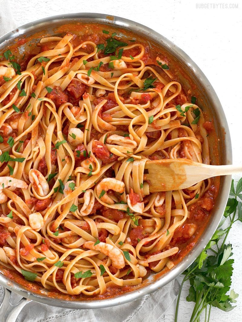 Spicy Seafood Pasta with Tomato Butter Sauce is a simple go-to weeknight dinner that can be made with pantry staples. BudgetBytes.com A skillet full of Spicy Seafood Pasta with Tomato Butter Sauce with a wooden fork twirled in the pasta and parsley on the side.