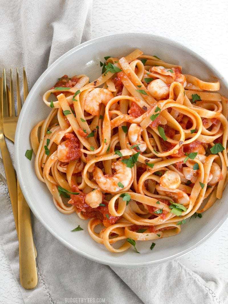 Spicy Seafood Pasta with Tomato Butter Sauce is a simple go-to weeknight dinner that can be made with pantry staples. BudgetBytes.com Close up overhead view of a bowl of Spicy Seafood Pasta with Tomato Butter Sauce with a gold fork and spoon on the side