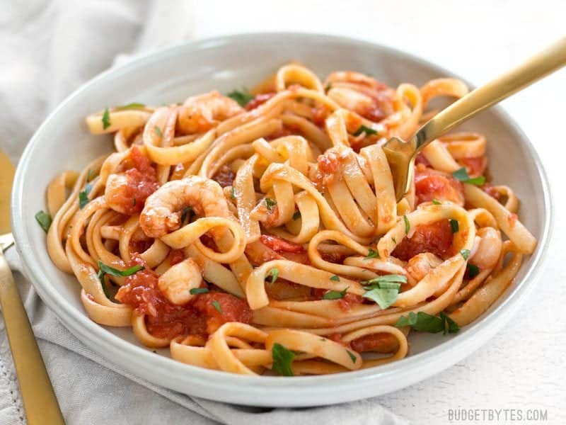 Spicy Seafood Pasta with Tomato Butter Sauce is a simple go-to weeknight dinner that can be made with pantry staples. BudgetBytes.com A bowl of Spicy Seafood Pasta with Tomato Butter Sauce viewed from the front, garnished with parsley, and a gold fork wound up in the pasta.