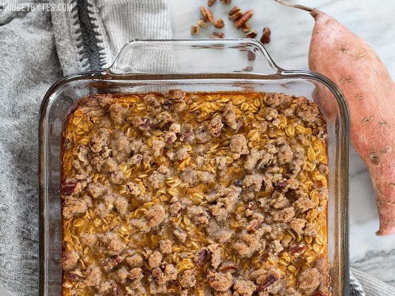 Sweet Potato Casserole Baked Oatmeal is a great way to have your favorite Thanksgiving side as a nutrient packed breakfast. BudgetBytes.com Finished Sweet Potato Casserole Baked Oatmeal in the glass casserole dish sitting on a napkin with a whole sweet potato and pecans on the side
