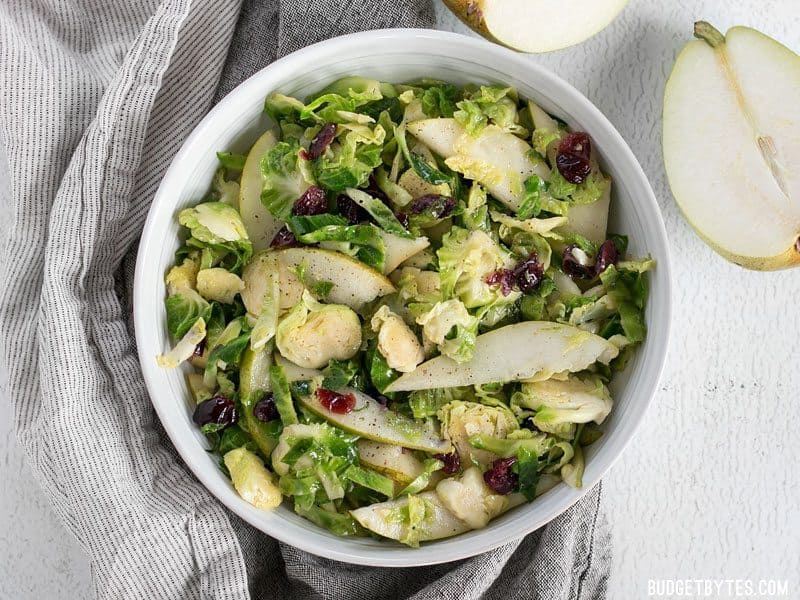 This Warm Brussels Sprouts and Pear Salad combines winter flavors in a warm and filling side dish. BudgetBytes.com Overhead shot of Warm Brussels Sprouts and Pear Salad in the bowl, a fresh pear on the side
