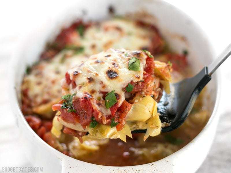 Baked Chicken with Artichokes and Tomatoes is an easy and flavorful weeknight dinner you can make with pantry staples. Budgetbytes.com A piece of Baked Chicken with Artichokes and Tomatoes being lifted out of the casserole dish with a spatula