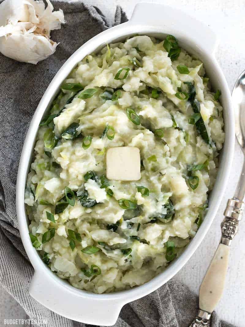 Colcannon is a simple Irish recipe that combines two hearty but inexpensive ingredients to make a delicious and filling side dish. BudgetBytes.com A large casserole dish full of Colcannon with a pat of butter melting on top and a head of garlic sitting on the side