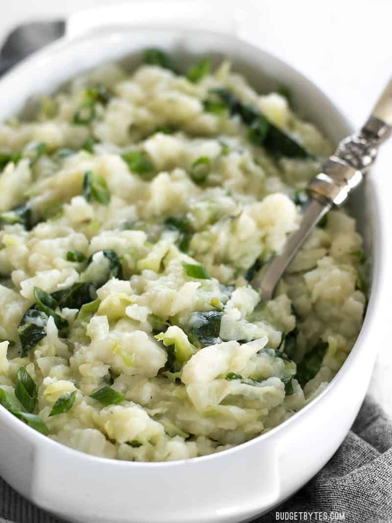 Colcannon is a simple Irish recipe that combines two hearty but inexpensive ingredients to make a delicious and filling side dish. BudgetBytes.com Side view of the casserole dish full of Colcannon, with a spoon stuck in the side.
