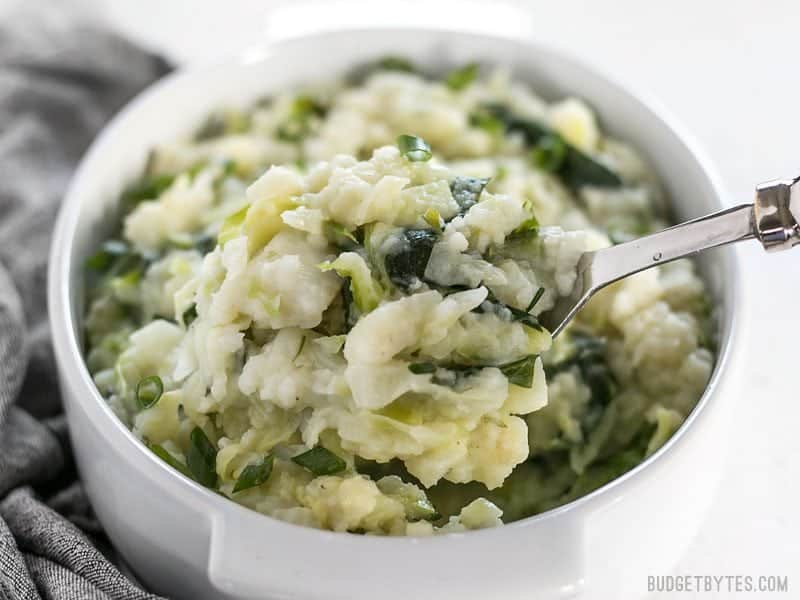 Colcannon is a simple Irish recipe that combines two hearty but inexpensive ingredients to make a delicious and filling side dish. BudgetBytes.com Front view of a casserole dish full of Colcannon with a big spoon lifting a heaping scoop.