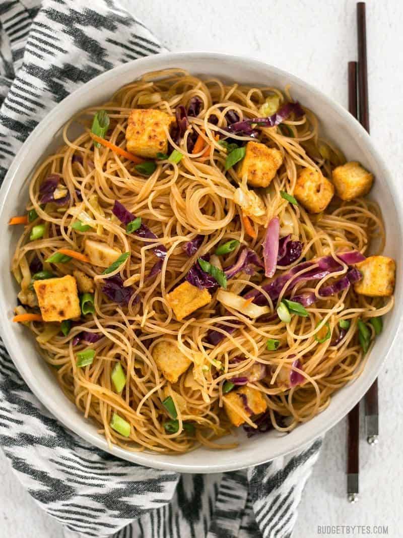 These Singapore Noodles with Crispy Tofu have a bold flavor and vibrant colors thanks to shredded vegetables and a bright curry sauce. BudgetBytes.com A big bowl of Singapore Noodles with Crispy Tofu on a patterned black and white napkin with chopsticks on the side