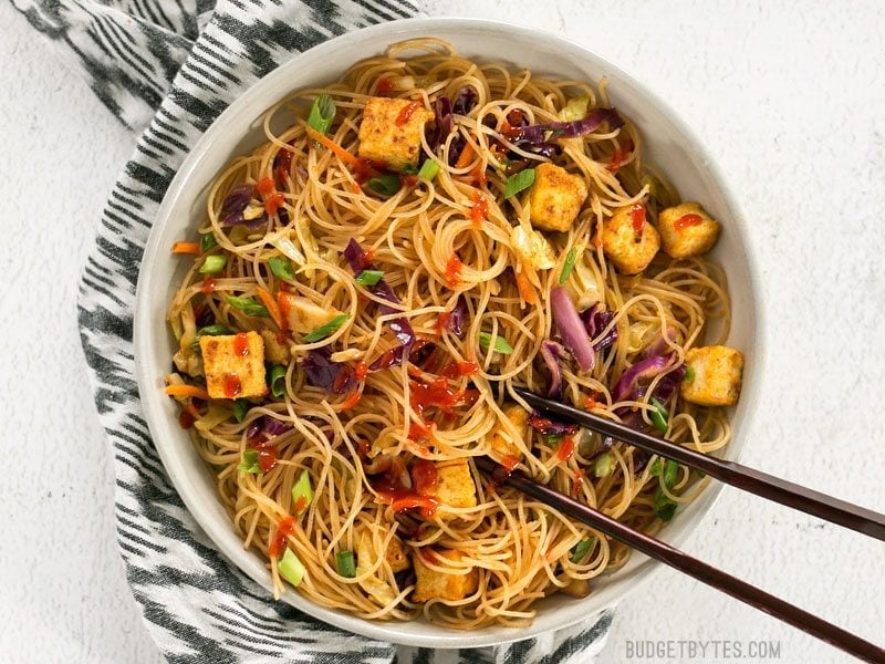 These Singapore Noodles with Crispy Tofu have a bold flavor and vibrant colors thanks to shredded vegetables and a bright curry sauce. BudgetBytes.com Overhead view of Singapore Noodles with Crispy Tofu garnished with sriracha, chopsticks in the bowl