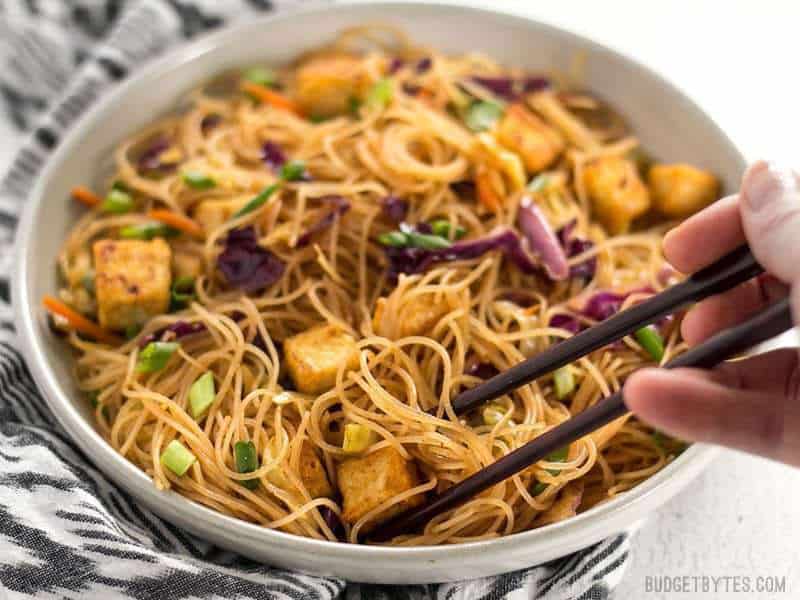 These Singapore Noodles with Crispy Tofu have a bold flavor and vibrant colors thanks to shredded vegetables and a bright curry sauce. BudgetBytes.com A bowl of Singapore Noodles with Crispy Tofu viewed from the front, chopsticks picking up a bunch of noodles