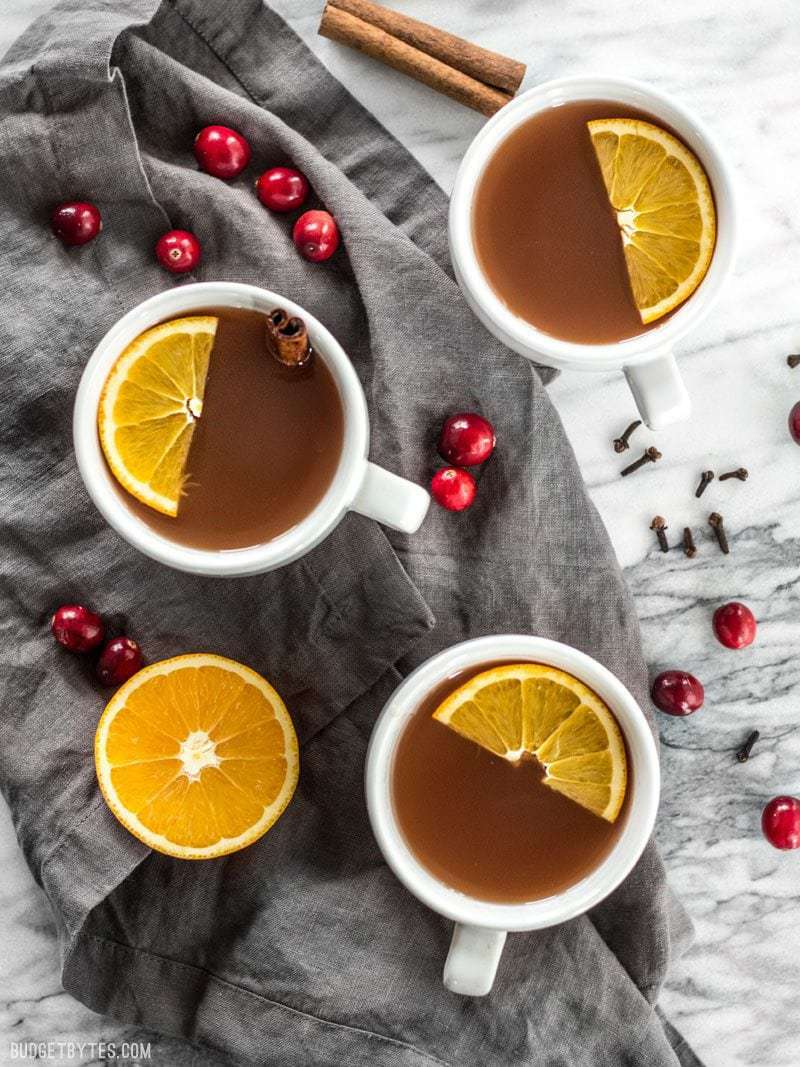 Slow Cooker Spiced Cranberry Apple Cider is an easy and festive drink for all your holiday party guests. BudgetBytes.com Three mugs of Slow Cooker Spiced Cranberry Apple Cider with fresh orange slices floating on top, and cranberries, cinnamon, and cloves sprinkled around