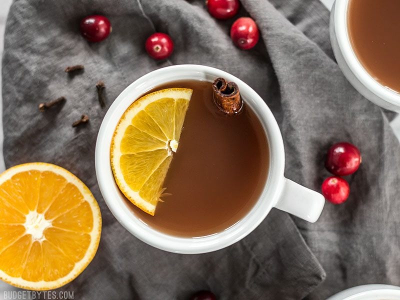 Slow Cooker Spiced Cranberry Apple Cider is an easy and festive drink for all your holiday party guests. BudgetBytes.com Close up of one mug full of Slow Cooker Spiced Cranberry Apple Cider with an orange slice and cinnamon stick