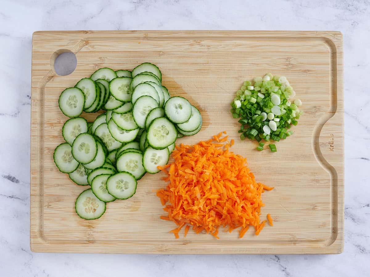 Sliced cucumber, shredded carrots, and diced green onions on a wooden cutting board.