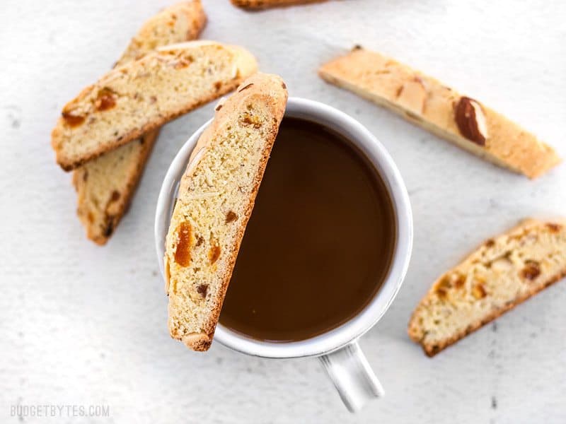 Fresh homemade Almond Apricot Biscotti are pennies a piece and can be stored in the freezer, ready for dunking at any time. BudgetBytes.com Almond Apricot Biscotti scattered around a cup of coffee, on balancing on the lip of the cup