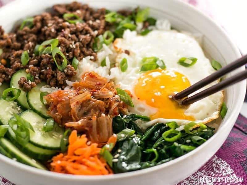 Bibimbap is the ultimate bowl meal with plenty of color, flavor, and texture to keep your taste buds happy and your stomach full. BudgetBytes.com Side view of a bowl of Bibimbap with the yolk being broken by a pair of chopsticks.