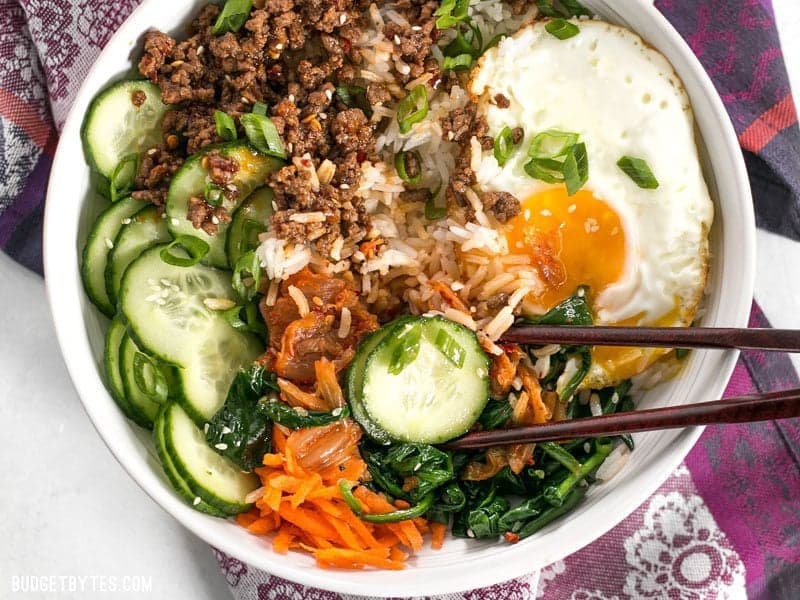 Bibimbap is the ultimate bowl meal with plenty of color, flavor, and texture to keep your taste buds happy and your stomach full. BudgetBytes.com Bird's eye view of a bowl of Bibimbap being eaten with chopsticks.