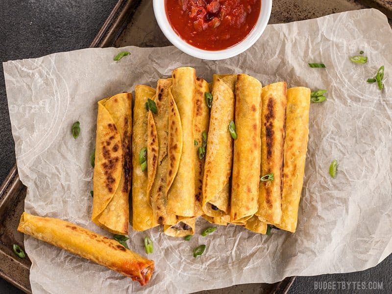 Creamy Black Bean Taquitos are an easy and tasty party treat for football games or just for fun! BudgetBytes.com A stack of Creamy Black Bean Taquitos on a piece of parchment, one has been dipped in salsa
