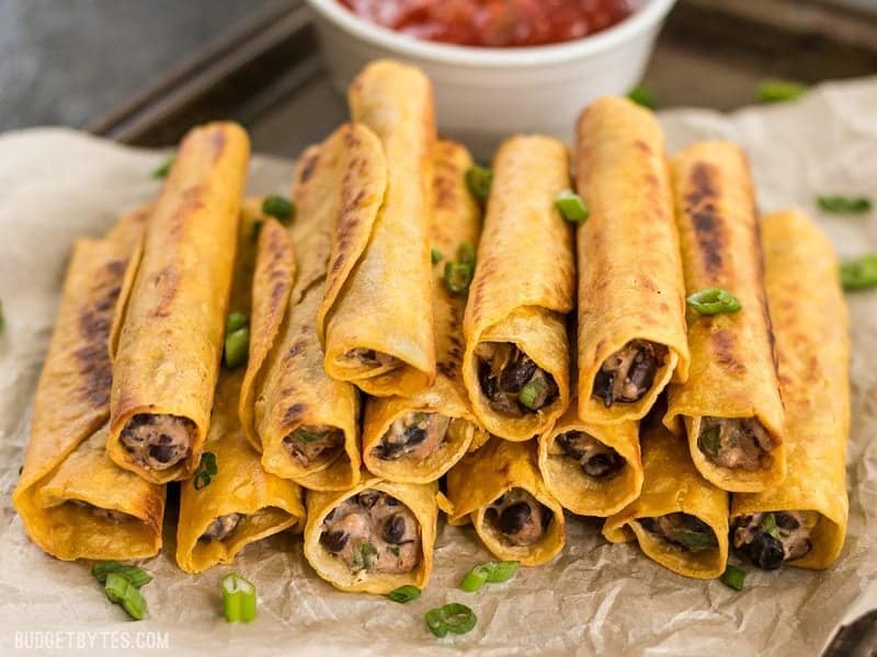 Creamy Black Bean Taquitos are an easy and tasty party treat for football games or just for fun! BudgetBytes.com Front view of a stack of Creamy Black Bean Taquitos with a bowl of salsa in the background