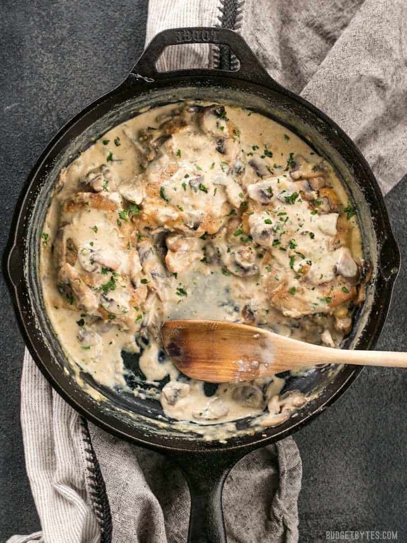 Simple pan sauces save the day in this quick and easy Creamy Garlic Mushroom Chicken! BudgetBytes.com Creamy Garlic Mushroom Chicken in a cast iron skillet, with a portion missing, a wooden spoon in its place