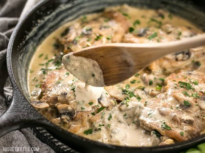 Simple pan sauces save the day in this quick and easy Creamy Garlic Mushroom Chicken! BudgetBytes.com Creamy Garlic sauce dripping off a wooden spoon onto the chicken thighs in the skillet