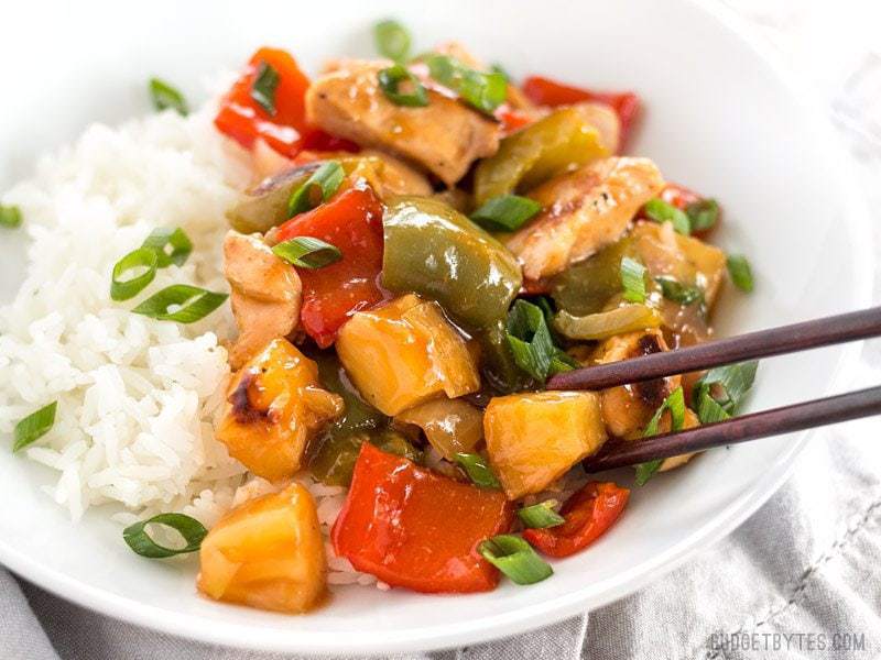 This oven baked Sheet Pan Sweet and Sour Chicken is a little more forgiving than a fast moving, high heat stir fry, making it great for beginners! BudgetBytes.com Close up of a plate of Sheet Pan Sweet and Sour Chicken over rice, a piece being picked up with chop sticks