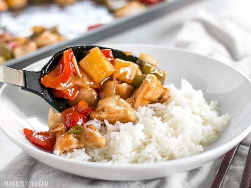 This oven baked Sheet Pan Sweet and Sour Chicken is a little more forgiving than a fast moving, high heat stir fry, making it great for beginners! BudgetBytes.com Sweet and Sour Chicken being spooned over a bowl of rice