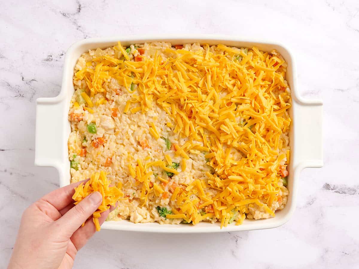 A hand sprinkling shredded cheese over a casserole dish of chicken rice filling