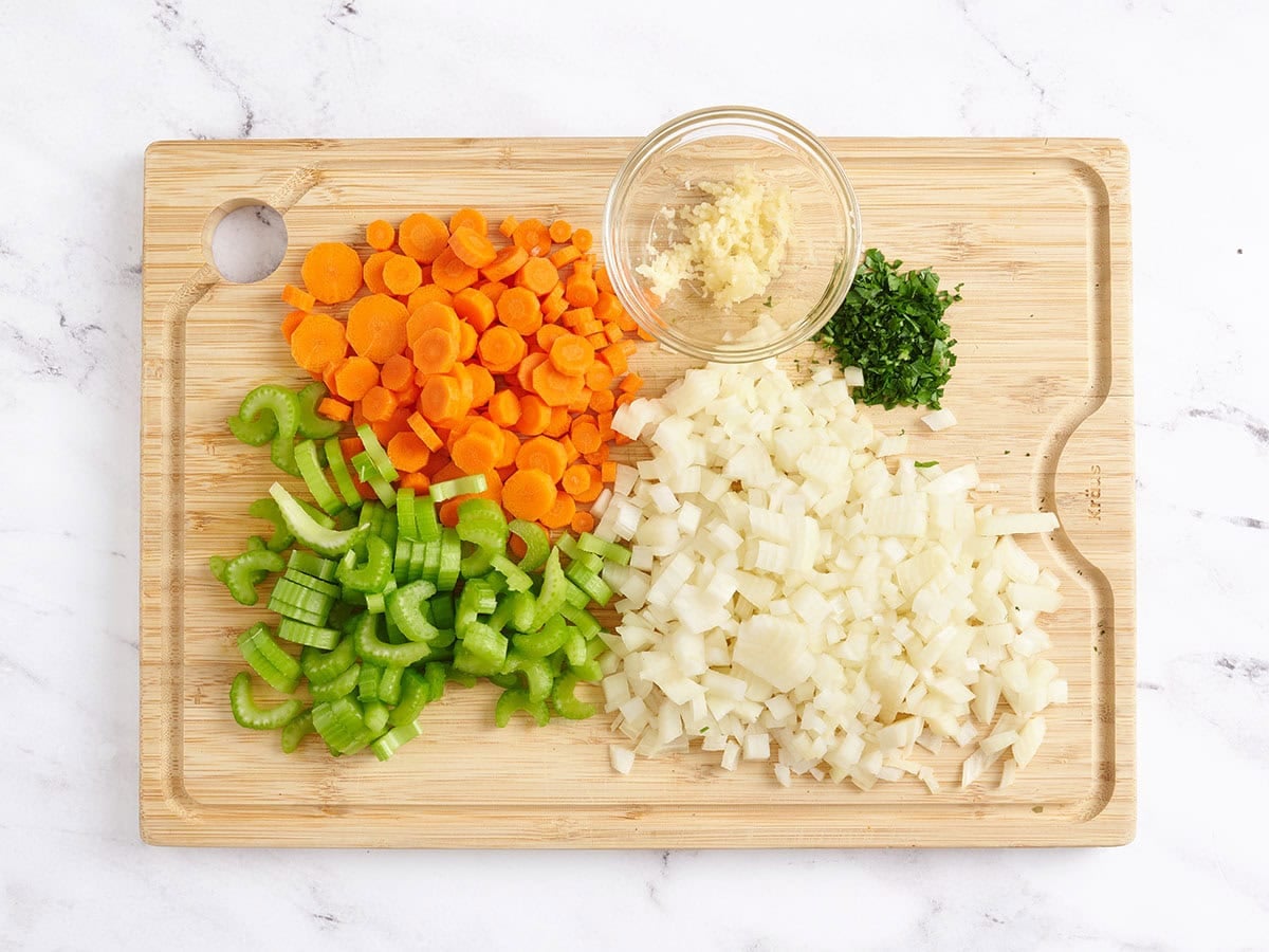 Diced carrots, celery, onion, minced parsley, and minced garlic on a wooden cutting board.