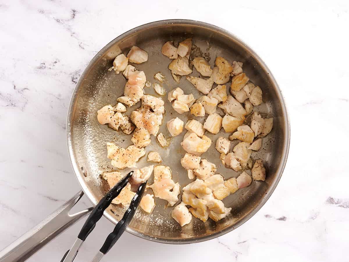 Diced chicken cooking in a skillet with tongs flipping a piece over